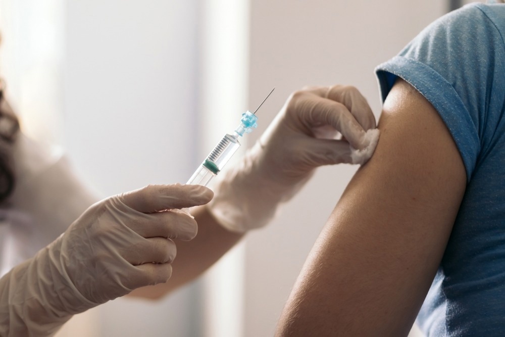 A healthcare professional wearing latex gloves administers a vaccine injection to a patient’s upper arm while using a cotton swab to prepare the site. The patient is wearing a short-sleeved blue shirt.