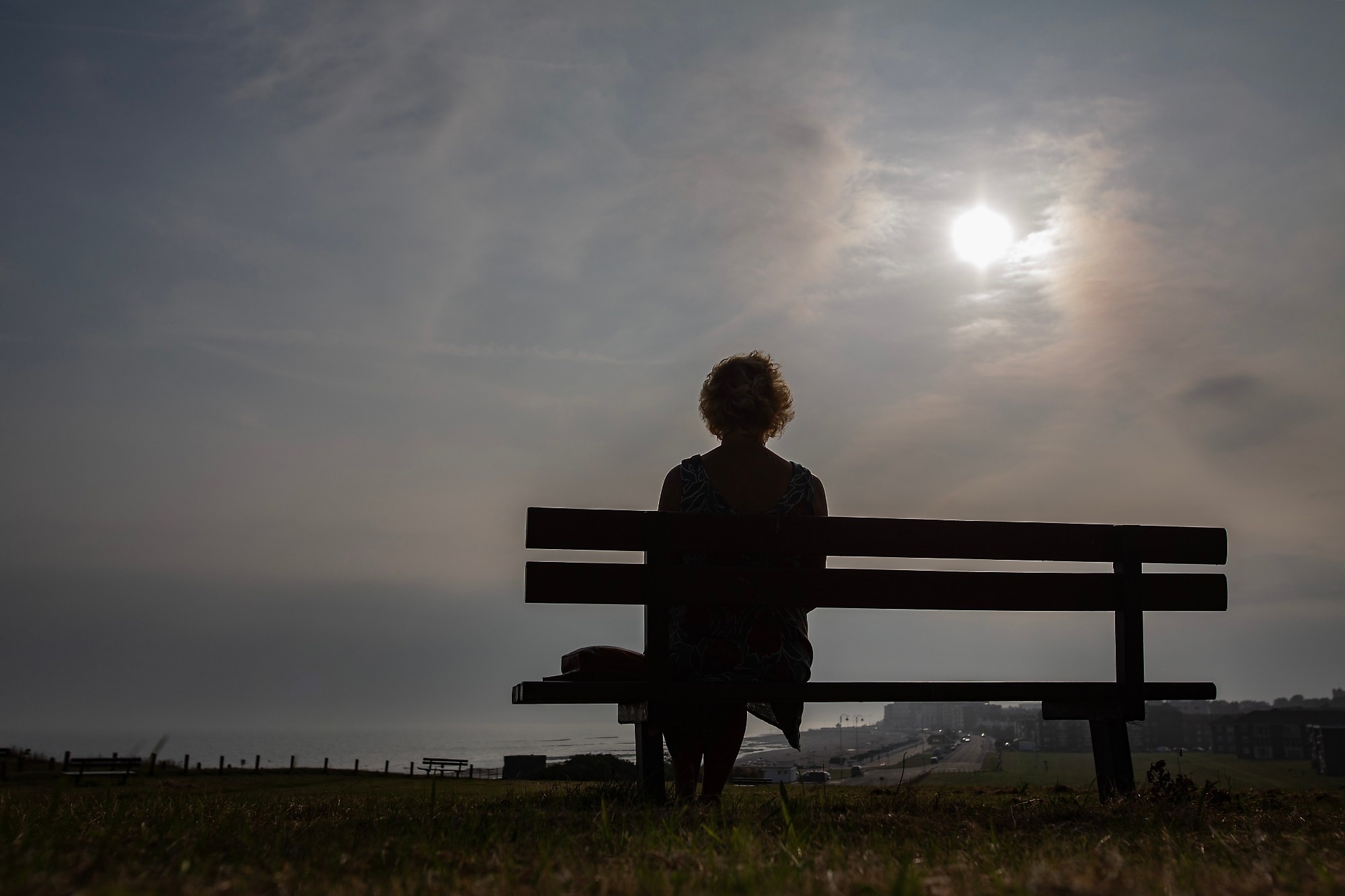 A lonely mature woman sits on a bench, on a hillside overlooking a coastal town.
