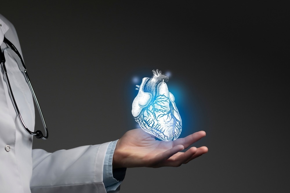 A doctor wearing a white coat and a stethoscope holds out their hand, presenting a digitally rendered, glowing blue anatomical heart floating above their palm. The heart is semi-transparent with detailed vein structures visible, symbolizing advanced medical technology and innovation in cardiology. The background is dark, emphasizing the illuminated heart.