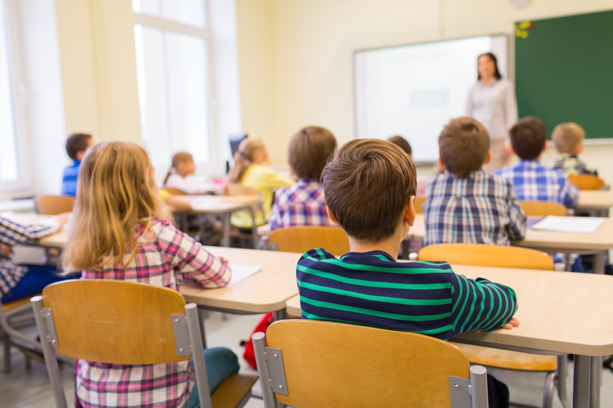group of school children sitting and listening to teacher in classroom from back